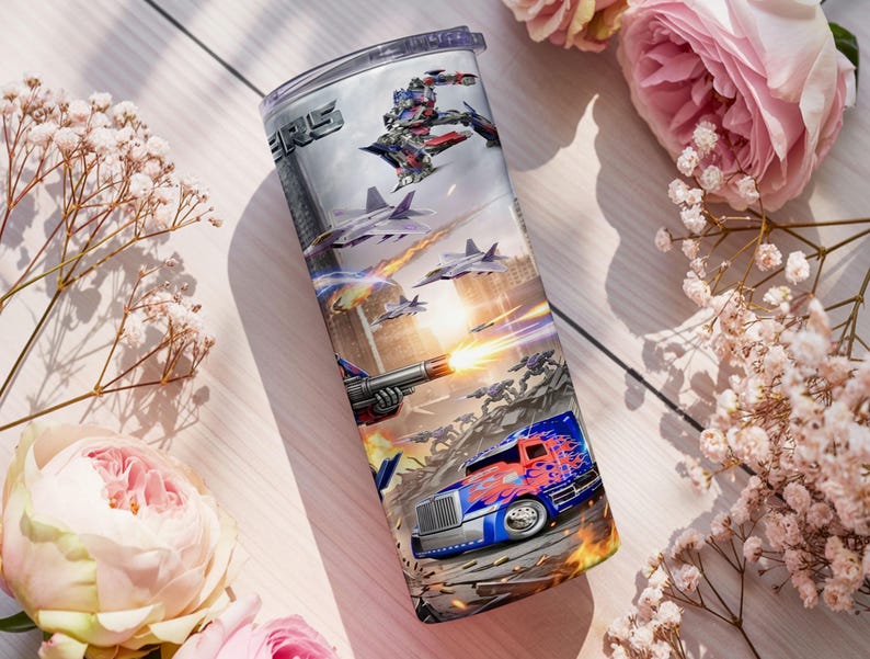 May include: A cylindrical metal tumbler featuring a full-wrap graphic print of a battle scene with a blue and red semi-truck, fighter jets, and a robotic character. The design includes fiery explosions and the partial text ERS in a metallic font.