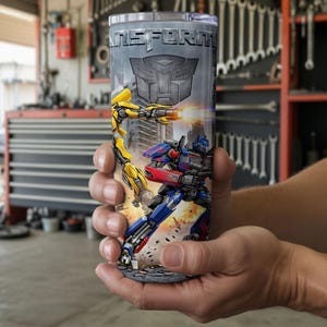 May include: A cylindrical stainless steel tumbler featuring a graphic print of two robotic characters in action, with the word TRANSFORM printed in bold metallic letters at the top and a large Autobot insignia centered below the text.