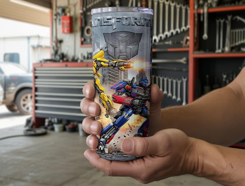 May include: A cylindrical stainless steel tumbler featuring a graphic print of two robotic characters in action, with the word TRANSFORM printed in bold metallic letters at the top and a large Autobot insignia centered below the text.