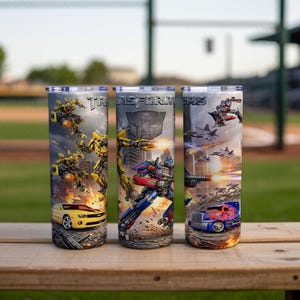 May include: Three cylindrical tumblers featuring Transformers graphics. The designs include Bumblebee in yellow car and robot forms, Optimus Prime in blue truck and robot forms, the Transformers logo, and action scenes with jets and explosions.