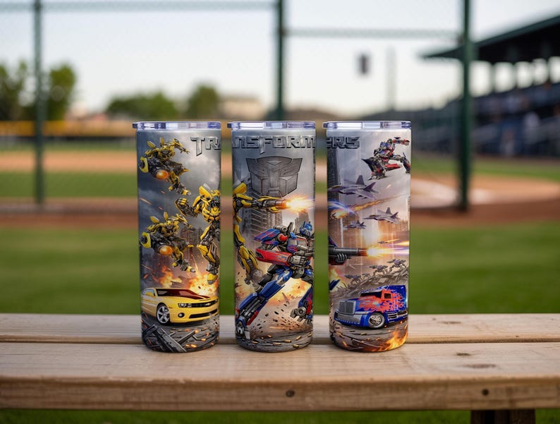 May include: Three cylindrical tumblers featuring Transformers graphics. The designs include Bumblebee in yellow car and robot forms, Optimus Prime in blue truck and robot forms, the Transformers logo, and action scenes with jets and explosions.