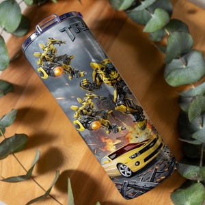 May include: A cylindrical stainless steel tumbler featuring a graphic print of the yellow robot character Bumblebee in various action poses, alongside a yellow sports car, set against a dark, smoky background with fiery orange explosions.