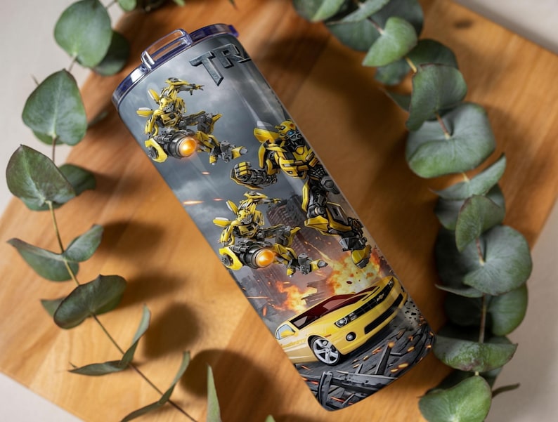 May include: A cylindrical stainless steel tumbler featuring a graphic print of the yellow robot character Bumblebee in various action poses, alongside a yellow sports car, set against a dark, smoky background with fiery orange explosions.