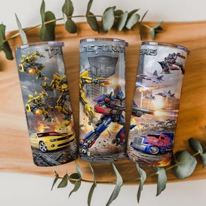 May include: Three cylindrical tumblers featuring Transformers graphics. The designs include yellow Bumblebee, blue and red Optimus Prime, and various vehicle forms like a yellow sports car and a blue semi-truck, all set against a battle scene background.