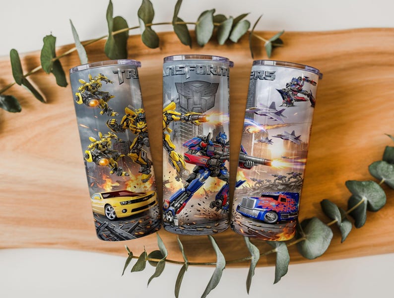 May include: Three cylindrical tumblers featuring Transformers graphics. The designs include yellow Bumblebee, blue and red Optimus Prime, and various vehicle forms like a yellow sports car and a blue semi-truck, all set against a battle scene background.