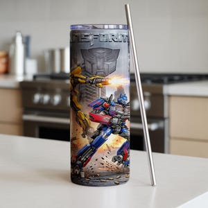 May include: A cylindrical stainless steel tumbler featuring a Transformers graphic with Optimus Prime and Bumblebee in action, the text TRANSFORMERS, and an Autobot logo. The tumbler includes a clear lid and a tall, thin silver metal straw.