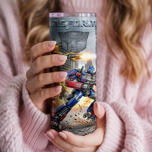 May include: A cylindrical stainless steel tumbler featuring a graphic print of Transformers characters, including a blue and red robot and a yellow robot firing a weapon. The word TRANSFORMERS is printed at the top with an Autobot logo below it.