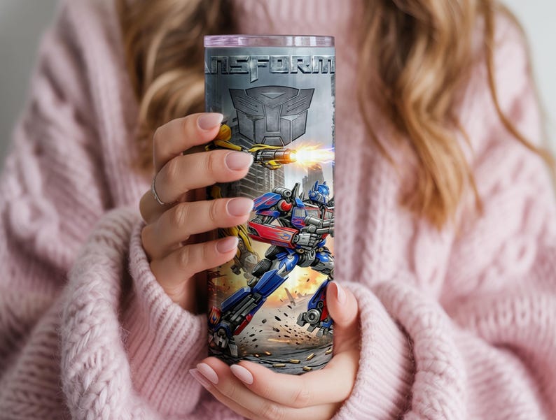 May include: A cylindrical stainless steel tumbler featuring a graphic print of Transformers characters, including a blue and red robot and a yellow robot firing a weapon. The word TRANSFORMERS is printed at the top with an Autobot logo below it.