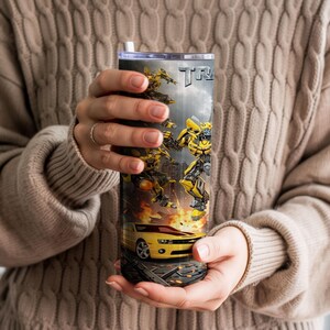 May include: A cylindrical stainless steel tumbler featuring a graphic print of yellow and black robotic characters in action, a yellow sports car, and the partial text "TRANS" against a dark, smoky background with orange flames at the base.