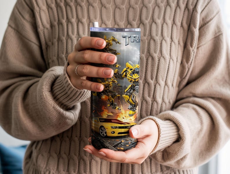 May include: A cylindrical stainless steel tumbler featuring a graphic print of yellow and black robotic characters in action, a yellow sports car, and the partial text "TRANS" against a dark, smoky background with orange flames at the base.
