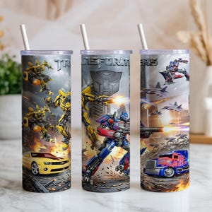 May include: Three cylindrical tumblers with white straws, featuring Transformers graphics. The designs include yellow and blue robot characters, a yellow sports car, a blue semi-truck, and the Transformers logo against a gray, action-filled background.