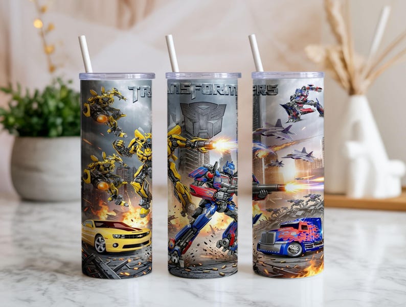May include: Three cylindrical tumblers with white straws, featuring Transformers graphics. The designs include yellow and blue robot characters, a yellow sports car, a blue semi-truck, and the Transformers logo against a gray, action-filled background.
