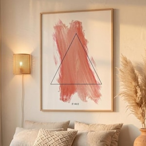 Könnte beinhalten: Ein gerahmter Kunstdruck mit einem abstrakten Pinselstrich in Korallen- und Pfirsichtönen und einem schwarzen Dreieck. Das Wort „FIRE“ ist darunter gedruckt. Das Kunstwerk hängt über einem Bett mit neutralfarbenen Kissen und einem gewebten Kissen.