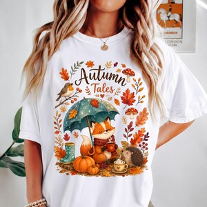 May include: A white t-shirt featuring a colorful graphic with the text "Autumn Tales". The design includes a fox holding a mug under a teal umbrella, a hedgehog, a robin, pumpkins, mushrooms, and orange and red autumn leaves.