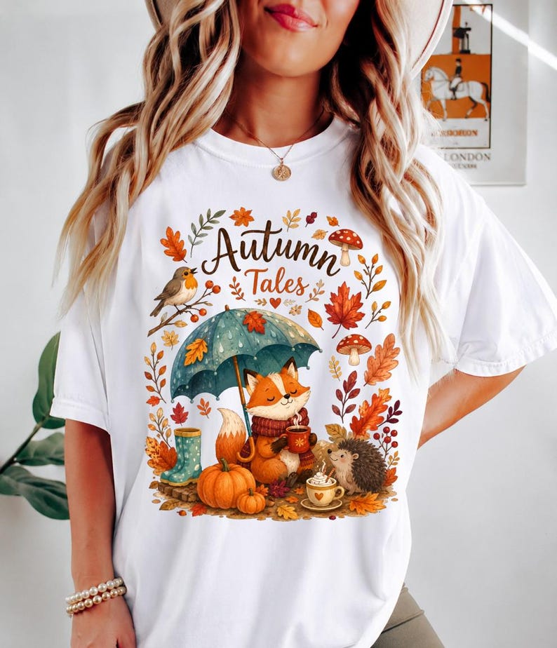 May include: A white t-shirt featuring a colorful graphic with the text "Autumn Tales". The design includes a fox holding a mug under a teal umbrella, a hedgehog, a robin, pumpkins, mushrooms, and orange and red autumn leaves.