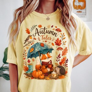 May include: A pale yellow t-shirt featuring a graphic print with the text "Autumn Tales". The design includes a fox holding a mug under a blue umbrella, a hedgehog, pumpkins, mushrooms, autumn leaves, a bird, and blue rain boots.