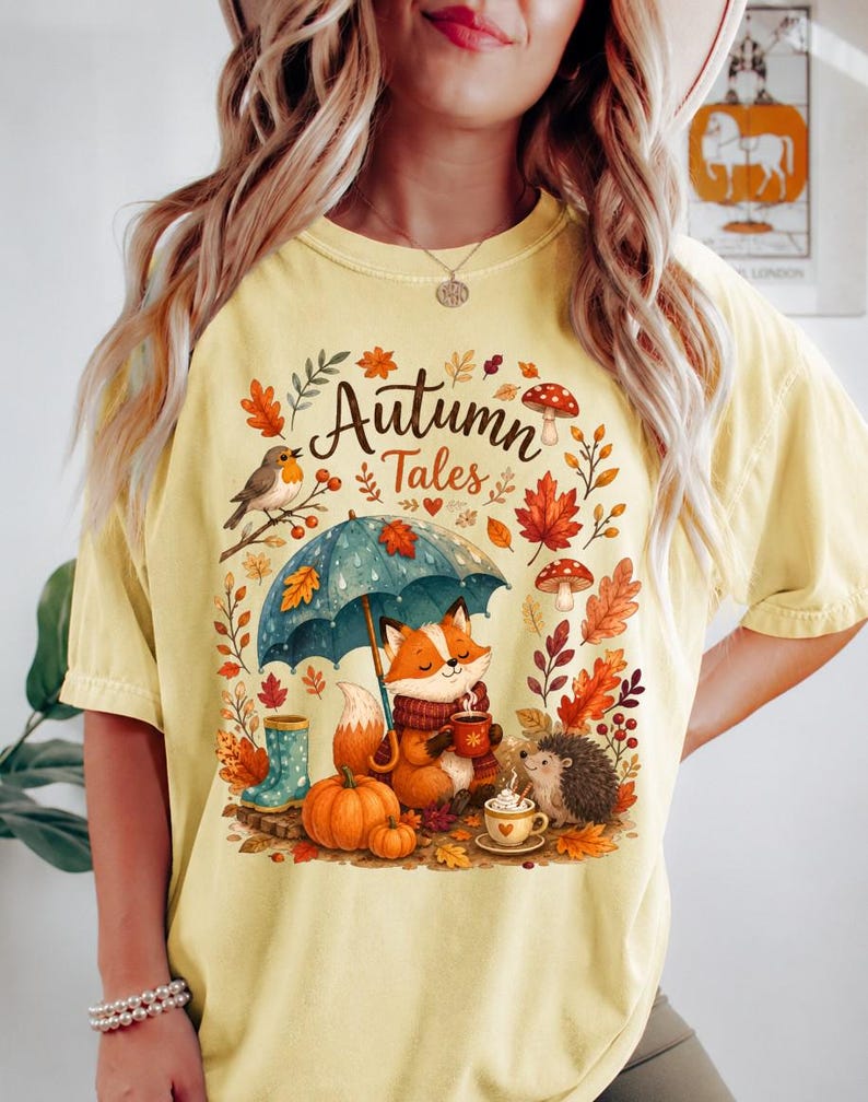 May include: A pale yellow t-shirt featuring a graphic print with the text "Autumn Tales". The design includes a fox holding a mug under a blue umbrella, a hedgehog, pumpkins, mushrooms, autumn leaves, a bird, and blue rain boots.