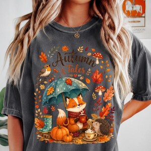 May include: A charcoal gray t-shirt featuring a graphic print with the text "Autumn Tales". The design includes a fox holding a mug under a teal umbrella, a hedgehog, a robin, pumpkins, mushrooms, and colorful fall leaves.