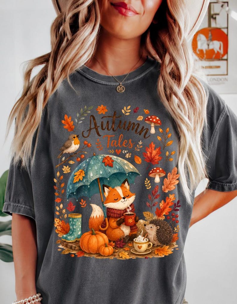 May include: A charcoal gray t-shirt featuring a graphic print with the text "Autumn Tales". The design includes a fox holding a mug under a teal umbrella, a hedgehog, a robin, pumpkins, mushrooms, and colorful fall leaves.