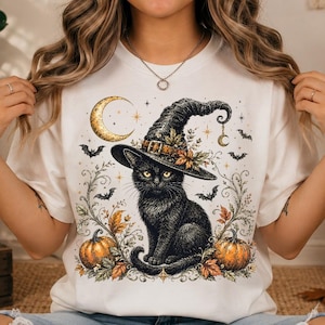 May include: A white t-shirt featuring a graphic print of a black cat wearing a witch hat, surrounded by orange pumpkins, autumn leaves, bats, and a golden crescent moon. The design includes intricate swirling vine details and small star accents.