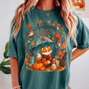 May include: A teal short-sleeve t-shirt featuring a graphic print with the text "Autumn Tales". The design includes a fox holding a mug under an umbrella, a hedgehog, pumpkins, mushrooms, a bird, and colorful fall leaves in shades of orange and red.