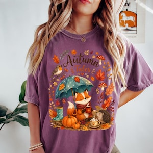 May include: A purple short-sleeve t-shirt featuring a graphic print with the text Autumn Tales. The design includes a fox holding a mug under a teal umbrella, a hedgehog, a pumpkin, a bird, mushrooms, and autumn leaves in orange and yellow.