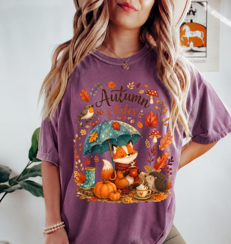 May include: A purple short-sleeve t-shirt featuring a graphic print with the text Autumn Tales. The design includes a fox holding a mug under a teal umbrella, a hedgehog, a pumpkin, a bird, mushrooms, and autumn leaves in orange and yellow.