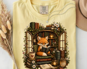 May include: A pale yellow t-shirt featuring a graphic of a fox wearing glasses and reading a book inside a library setting. The illustration includes shelves of books, a steaming mug with a heart, a lantern, and white flowers. The shirt has a Comfort Colors label.