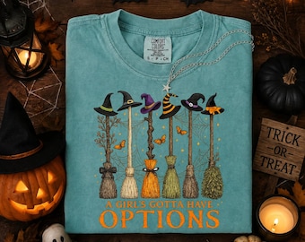 May include: A teal short-sleeve t-shirt featuring a graphic of six different witch brooms with colorful hats and butterflies. The text A GIRL'S GOTTA HAVE OPTIONS is printed in orange below the brooms. The shirt has a Comfort Colors brand label.