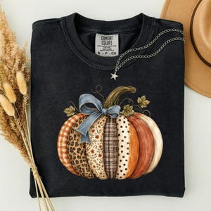 May include: A black crewneck t-shirt featuring a large pumpkin graphic composed of various patterns including plaid, leopard print, floral, and polka dots, accented with a blue ribbon bow. The shirt has a Comfort Colors label at the inner neckline.