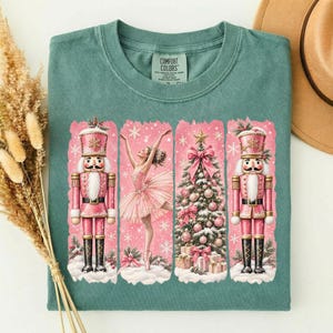 May include: A sage green t-shirt featuring a graphic print of two pink nutcrackers, a ballerina in a pink tutu, and a decorated Christmas tree. The design includes white snowflakes and the text COMFORT COLORS on the inner neck label.