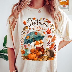 May include: A cream-colored t-shirt featuring a graphic print with the text "Autumn Tales" in a script font. The illustration depicts a fox holding a mug under a blue umbrella, a hedgehog, pumpkins, mushrooms, a bird, and autumn leaves.