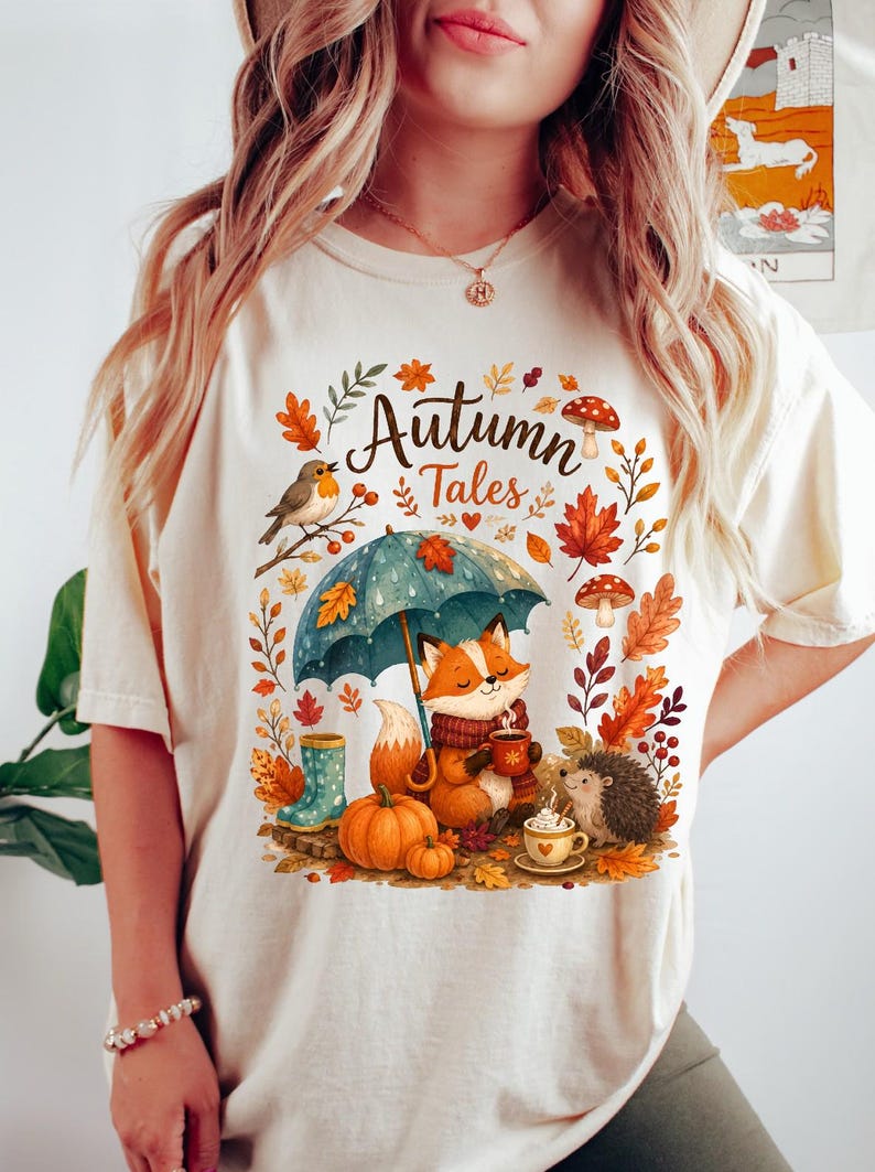 May include: A cream-colored t-shirt featuring a graphic print with the text "Autumn Tales" in a script font. The illustration depicts a fox holding a mug under a blue umbrella, a hedgehog, pumpkins, mushrooms, a bird, and autumn leaves.