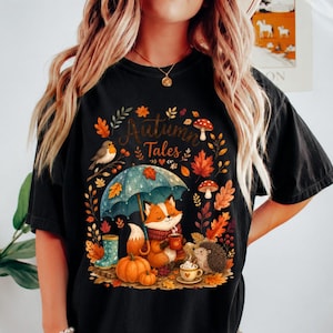 May include: A black t-shirt featuring a graphic print with the text Autumn Tales. The design includes a fox holding a mug under a blue umbrella, a hedgehog, a bird, pumpkins, mushrooms, and autumn leaves in shades of orange, red, and yellow.