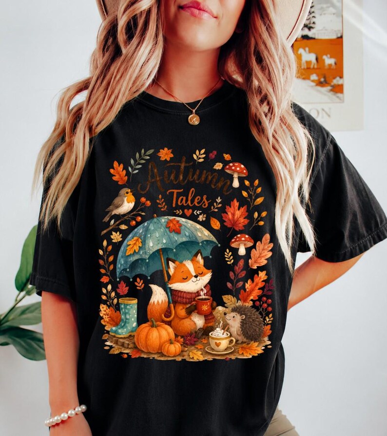 May include: A black t-shirt featuring a graphic print with the text Autumn Tales. The design includes a fox holding a mug under a blue umbrella, a hedgehog, a bird, pumpkins, mushrooms, and autumn leaves in shades of orange, red, and yellow.