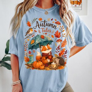 May include: A light blue t-shirt featuring a graphic print with the text "Autumn Tales". The illustration depicts a fox holding a mug under a teal umbrella, a hedgehog, pumpkins, mushrooms, and autumn leaves in shades of orange and brown.