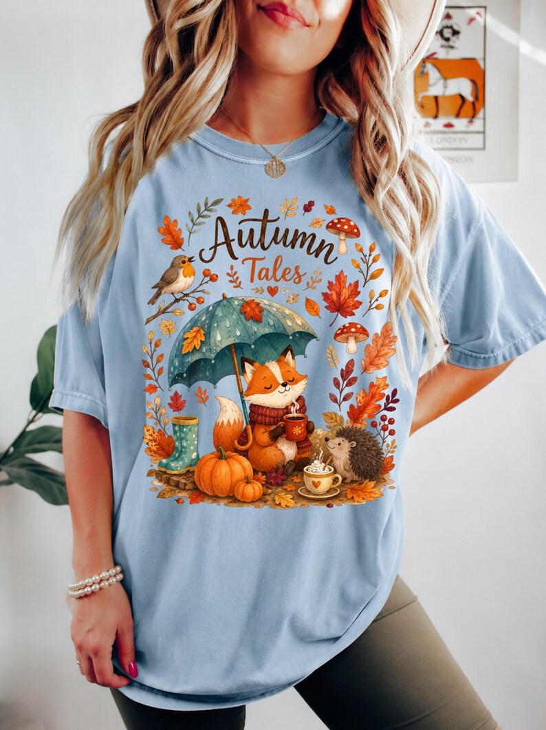 May include: A light blue t-shirt featuring a graphic print with the text "Autumn Tales". The illustration depicts a fox holding a mug under a teal umbrella, a hedgehog, pumpkins, mushrooms, and autumn leaves in shades of orange and brown.