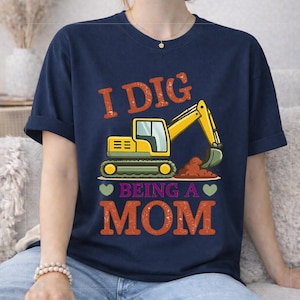 May include: Navy blue t-shirt with the text "I DIG BEING A MOM" in orange and green. The graphic features a yellow excavator digging brown dirt. Two green hearts are on either side of the words "BEING A".