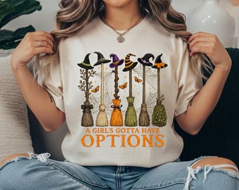 May include: A cream-colored t-shirt featuring a graphic of six different witch brooms with various hats, spiderwebs, and butterflies. Below the illustration, the text reads A GIRL'S GOTTA HAVE OPTIONS in bold orange capital letters.