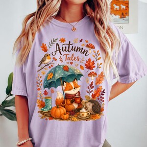 May include: A light purple t-shirt featuring a graphic print with the text "Autumn Tales". The design includes a fox holding a mug, a hedgehog, a pumpkin, a teal umbrella, mushrooms, and autumn leaves in shades of orange, red, and brown.