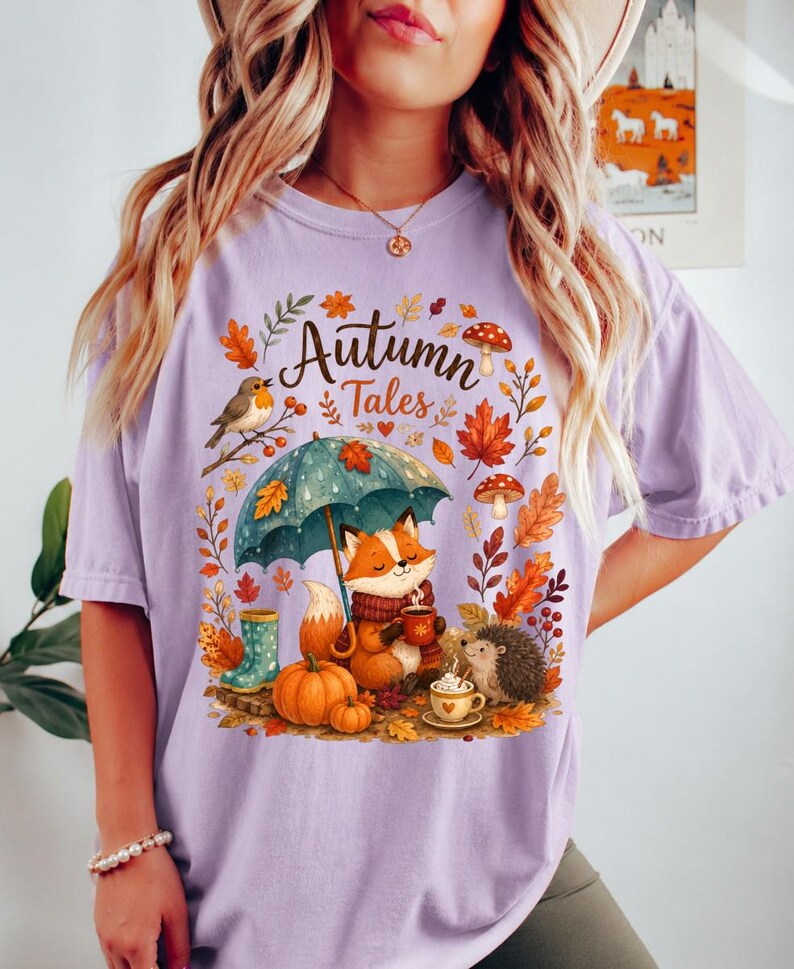 May include: A light purple t-shirt featuring a graphic print with the text "Autumn Tales". The design includes a fox holding a mug, a hedgehog, a pumpkin, a teal umbrella, mushrooms, and autumn leaves in shades of orange, red, and brown.