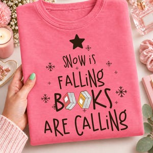 May include: A bright pink t-shirt featuring a black graphic design with a star at the top, snowflake icons, and the text "SNOW IS FALLING BOOKS ARE CALLING" with two small book illustrations integrated into the word "BOOKS" in the center.