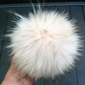 May include: A fluffy, round, cream-colored fur pom-pom with long, wispy hairs. The pom-pom has a light brown base with darker tips, creating a textured appearance. This accessory is likely for hats or other apparel.