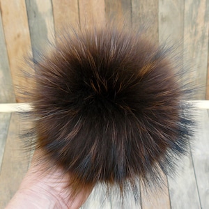 May include: A fluffy, round, brown fur pompom with dark tips. The pompom is held in a hand, showcasing its texture and color variations. The fur appears soft and dense, with a mix of light and dark brown shades.