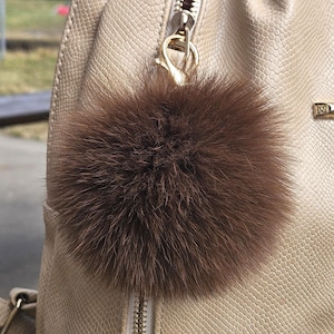 May include: A brown fur pom-pom attached to a beige backpack with a gold zip. The pom-pom is round and fluffy, adding a decorative element to the bag. The backpack has a textured, snake-skin-like pattern.