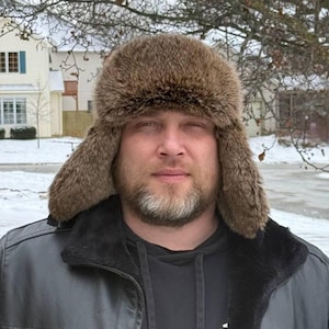 May include: A person wearing a brown fur hat with ear flaps and a black leather jacket with a black fleece collar. The background shows a snowy residential area with houses and trees.
