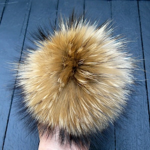 May include: A fluffy, round, faux fur pom-pom in shades of tan, brown, and black. The pom-pom has a dense, textured appearance with long, spiky strands. The item is likely for hats, scarves, or other accessories.