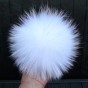 May include: A fluffy, round, white fur pom-pom. The pom-pom is held in a hand and appears to be made of soft, long fur fibers. The background is a dark, textured surface.