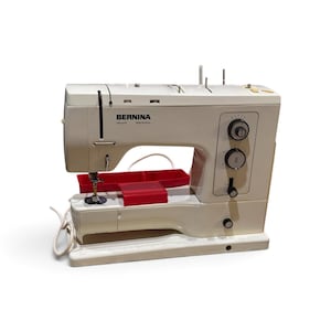 Op de afbeelding: Een crèmekleurige Bernina record electronic naaimachine met een witte behuizing, zwarte bedieningsknoppen aan de rechterkant en een rood plastic accessoirebakje op de basis. De machine is voorzien van een naald, naaivoet en stroomkabel.