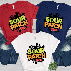 May include: Three t-shirts in red, white, and navy blue, each featuring the "Sour Patch Kids" logo. The logo includes the text "Sour Patch Kids" in a mix of green, orange, and red, with a cluster of black candy figures around it.